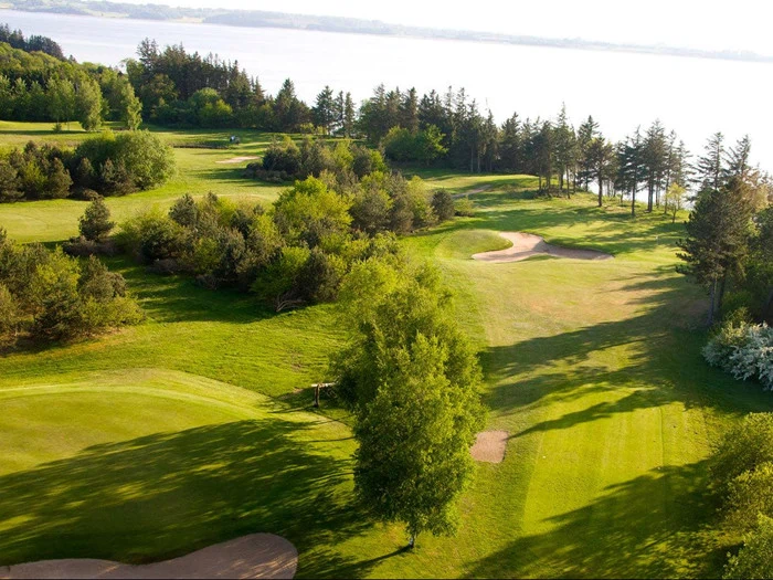 Luftfoto af en frodig grøn golfbane med sandbunkere, træer og en sø i baggrunden i stærkt sollys.