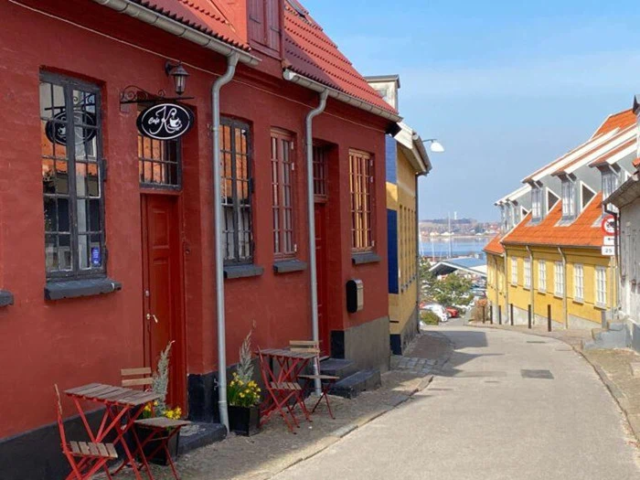 En smal brostensbelagt gade med farverige bygninger, udendørs caféborde med røde stole og udsigt til vand i det fjerne under en blå himmel.