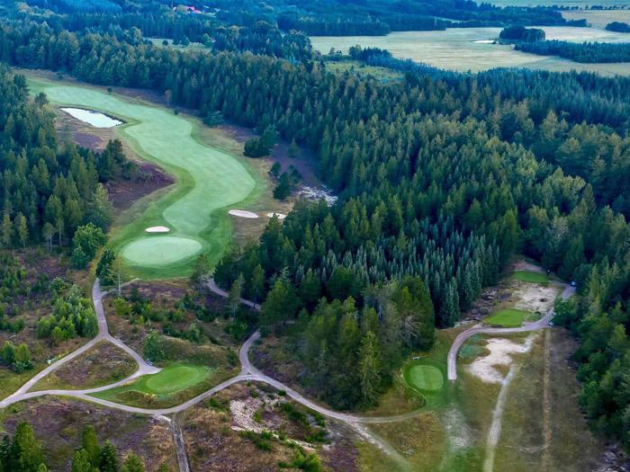 Luftfoto af en golfbane omgivet af tæt skov med grønne fairways, sandbunkers og snoede vognbaner.