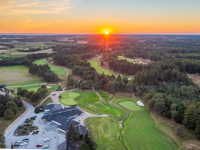 Luftfoto af en golfbane ved solnedgang med grønne fairways, sandfælder, træer og et klubhus i forgrunden under en orange himmel.