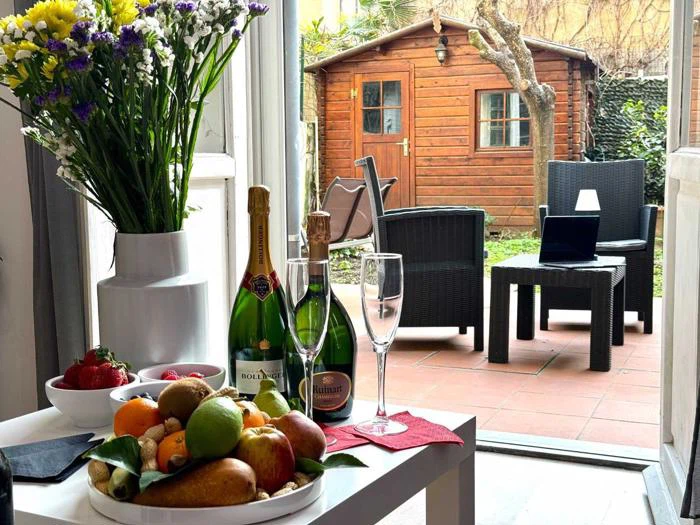 Et bord med frugt, blomster, to flasker champagne og glas står ved et vindue med udsigt til en terrasse med kurvestole, en bærbar computer og et træskur i en grøn have.