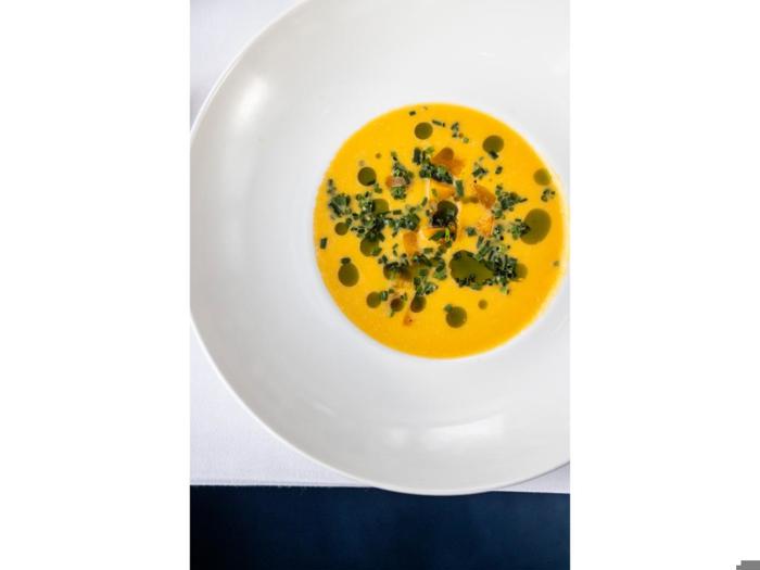 En hvid skål fyldt med lys orange suppe, pyntet med grønne oliedråber og finthakkede krydderurter, placeret på en hvid dug.