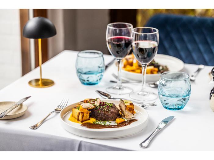 Et dækket spisebord med to tallerkener med bøf, kartofler, grøntsager, to glas rødvin, blå vandglas, sølvtøj og en sort lampe på en hvid dug.