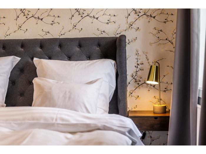 En pænt redt seng med hvidt sengetøj, et tuftet gråt hovedgærde, blomstret tapet og en guld sengelampe på et natbord af træ.