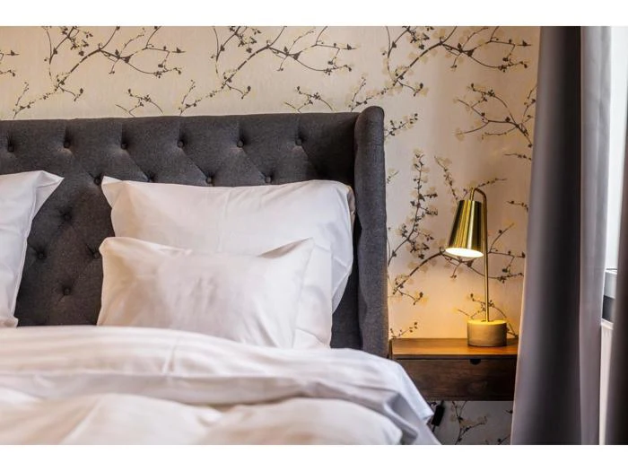 En pænt redt seng med hvidt sengetøj, et tuftet gråt hovedgærde, blomstret tapet og en guld sengelampe på et natbord af træ.