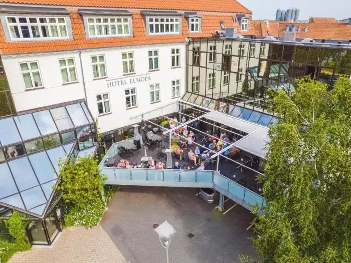 Luftfoto af mennesker, der spiser på en glasoverdækket terrasse på Hotel Europa, en hvid bygning med orange tagsten og store vinduer.