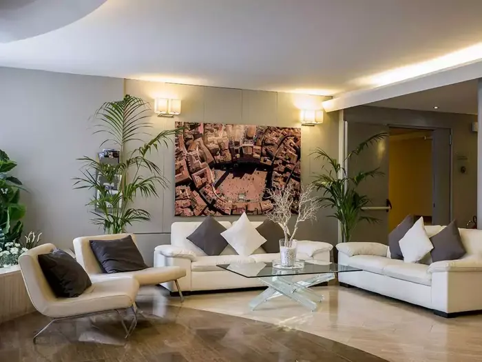 Moderne stue med hvide sofaer, sofabord i glas, grønne planter og et stort luftfoto af byen på væggen. Neutrale toner og blød belysning skaber en hyggelig, elegant atmosfære.