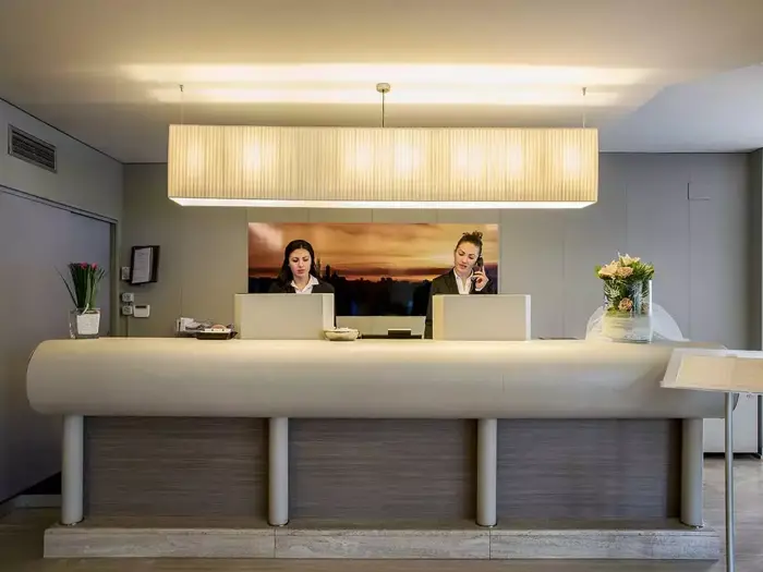 To receptionister står bag en moderne hotelreception og arbejder ved computere. En stor lampe hænger ovenover, og blomster pynter på disken.