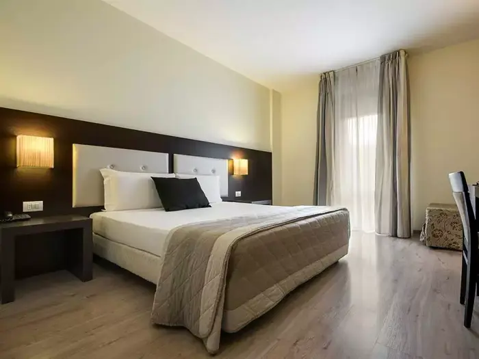 Moderne hotelværelse med en stor seng, to natborde, væghængte lamper, gennemsigtige gardiner over et vindue og lyst trægulv.