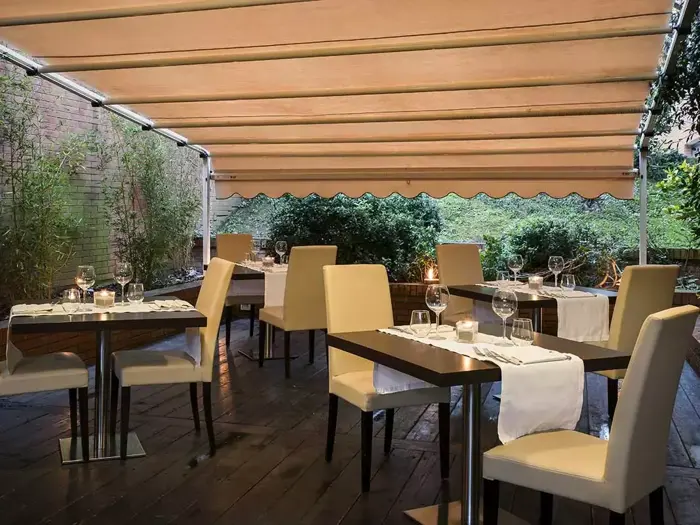 Udendørs restaurantterrasse med beige stole og borde til spisning, hvide duge, vinglas, stearinlys og en beige udtrækkelig markise; grønne planter i baggrunden.