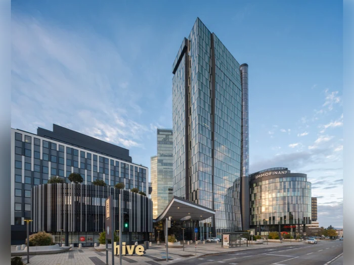 En moderne bygade med høje kontorbygninger i glas, bl.a. en med navnet IMMOFINANZ og en anden med et skilt, hvor der står bikube, under en blå himmel med spredte skyer.