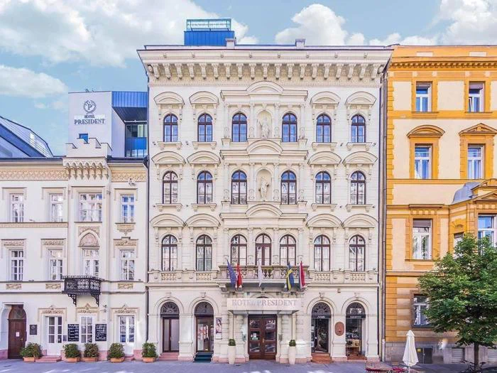 Elegant hvid historisk bygning med udsmykkede detaljer, mærket Hotel President, flankeret af bygninger i samme stil under en delvist overskyet himmel. Flere nationale flag hænger over indgangen.