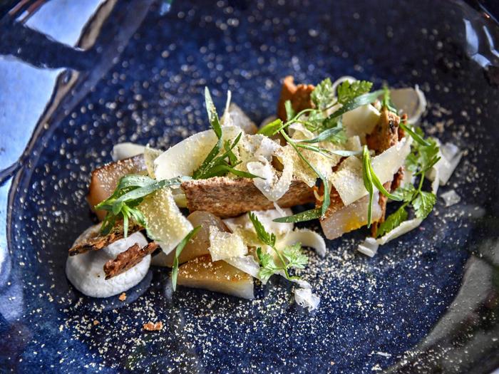 En gourmetret med skiveskåret ost, friske krydderurter, pærestykker og sprødt brød på en mørkeblå tallerken, pyntet med fint hvidt pulver.