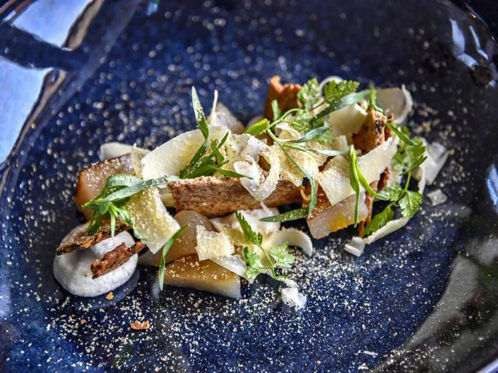 En gourmetret med tynde spåner af ost, friske krydderurter, sprødt brød og skiveskårne grøntsager kunstfærdigt anrettet på en mørkeblå keramiktallerken.