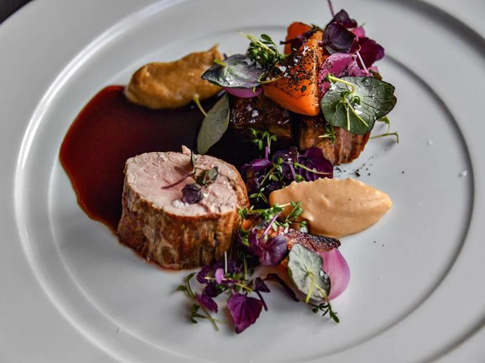 En gourmetret med kød i skiver, cremede puréer, grønt, spiselige blomster og en mørk sauce, kunstfærdigt anrettet på en hvid tallerken.