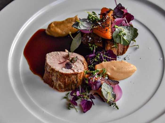 En gourmetret med skiveskåret kød, to saucer og pyntet med mikrogrønt, krydderurter og spiselige blomster på en hvid tallerken.