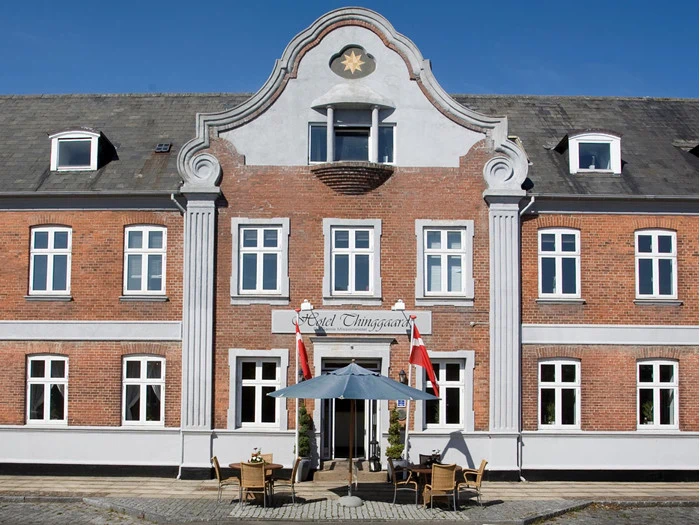 Et hotel i røde mursten med hvide kanter, høje vinduer, en dekorativ gavl og udendørs siddepladser under en blå parasol med to danske flag ved indgangen.