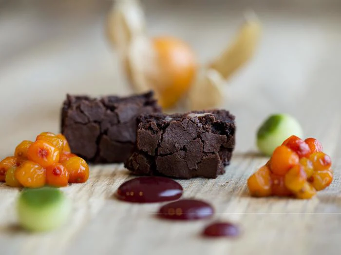 To stykker chokoladebrownie på en træoverflade, omgivet af bær, frugtpuré og saucedråber, med en sløret frugt i baggrunden.