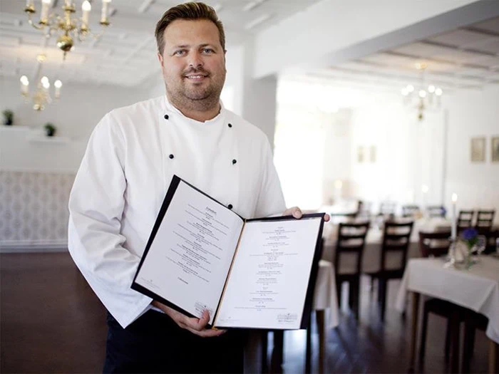 En kok i hvid uniform står i en restaurantspisestue, smiler og holder en åben menu op mod kameraet.