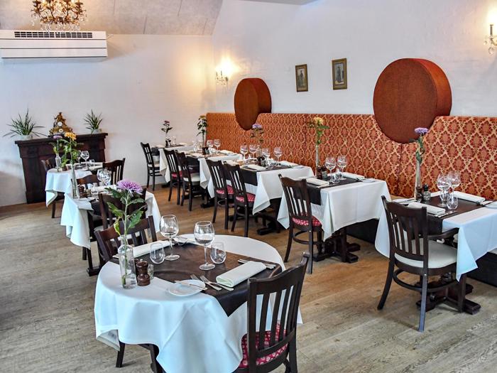 Elegant restaurantinteriør med spiseborde, hvide duge, vinglas, mørke træstole og dekorativ rød vægbeklædning. Rummet er lyst og indbydende.