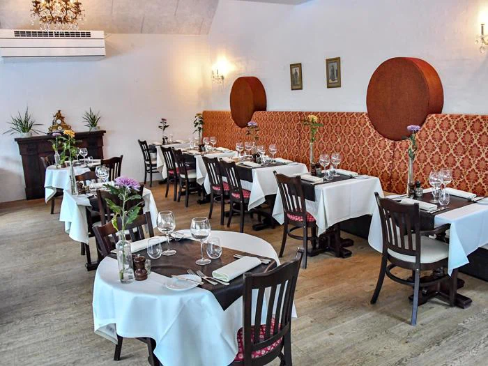 Elegant restaurantinteriør med spiseborde, hvide duge, vinglas, mørke træstole og dekorativ rød vægbeklædning. Rummet er lyst og indbydende.