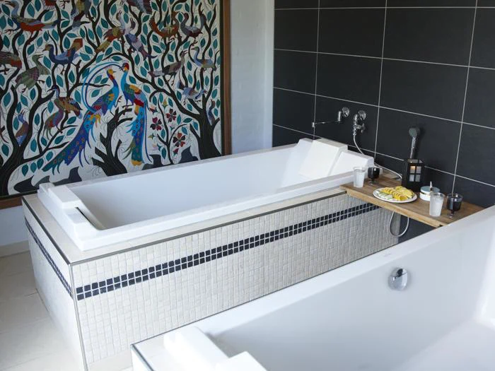 Et moderne badeværelse med to hvide badekar, en sort flisebelagt væg og et farverigt vægmaleri af fugle og træer. Det ene badekar har en træbakke med snacks, drikkevarer, et håndklæde og en lanterne.