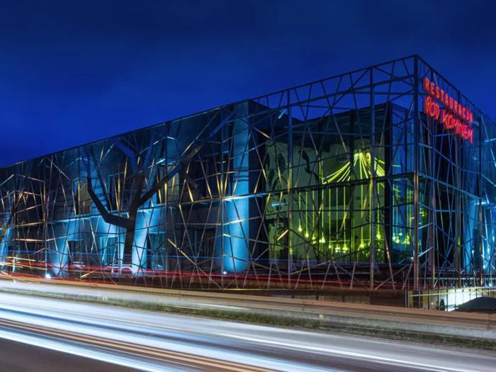 En moderne bygning med blå belysning, geometriske metalrammer og et glødende gult indre med lysspor fra forbipasserende biler på vejen foran.