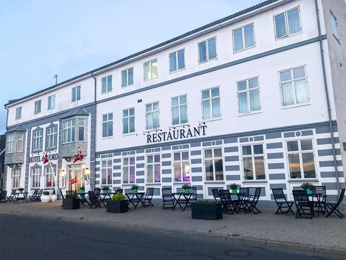 En hvid treetagers bygning med RESTAURANT- og HOTEL NORD-skilte, udeservering, potteplanter og danske flag ved indgangen.