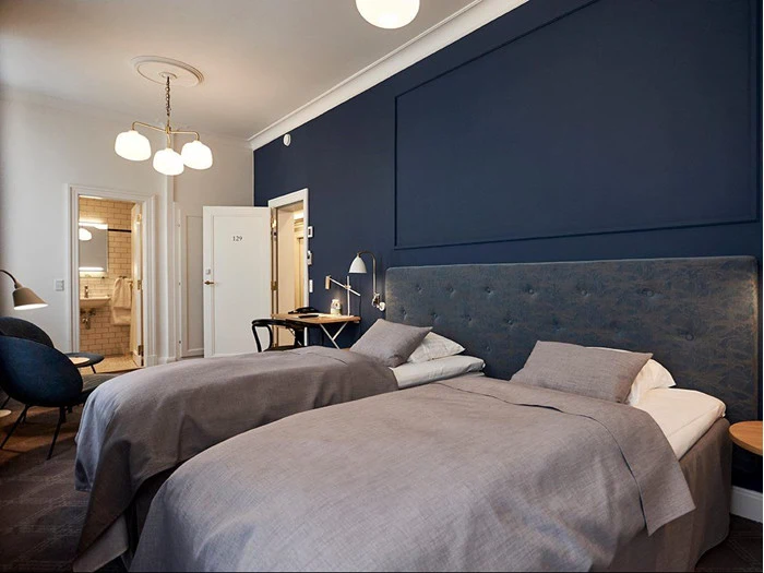 Moderne hotelværelse med to enkeltsenge, gråt sengetøj, en mørkeblå accentvæg, et skrivebord, stole og en åben dør, der fører til et badeværelse med flisebelagte vægge.