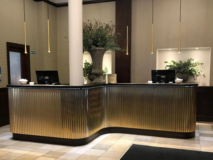 En stilfuld hotelreception med gyldne detaljer, to computerskærme, potteplanter og hængende pendellamper over.