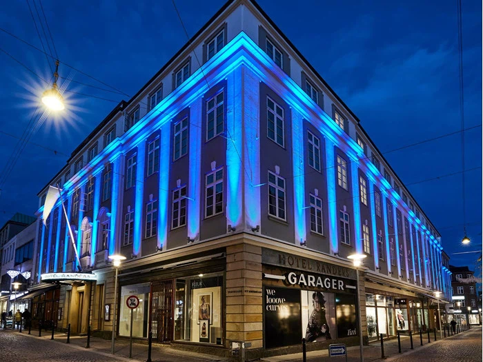 En hjørnebygning om natten med blå lys, der oplyser den øverste facade; den nederste etage har butiksvinduer og skilte, og gaden er stille og tom.