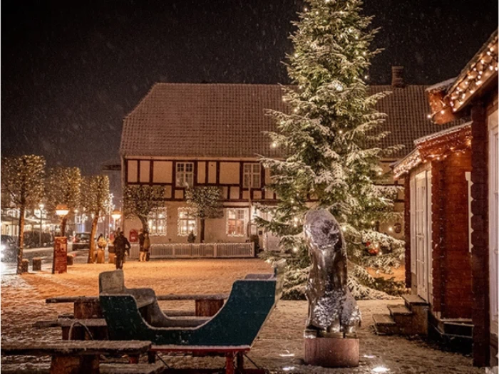 Sneen falder blidt over et festligt bytorv om natten med et oplyst juletræ, en kane og varmt oplyste bygninger pyntet med julelys.