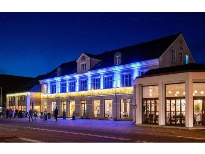 En stor bygning med øvre og nedre vinduer er udsmykket med blå og gule lys om natten. Flere mennesker går forbi på fortovet foran.