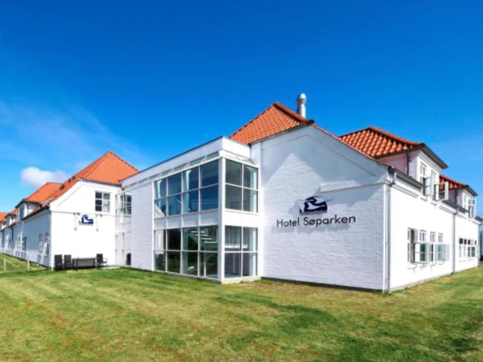 En hvid bygning med rødt tegltag og store vinduer med navnet Hotel Søparken ligger på en græsplæne under en klar blå himmel.