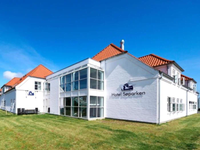 En hvid hotelbygning med rødt tegltag og store vinduer med navnet Hotel Søparken mod en lyseblå himmel og grønt græs.