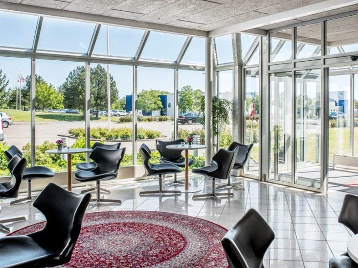 Moderne venteområde med sorte stole, små borde og et rødt mønstret tæppe, omgivet af store vinduer, der viser træer, biler og en lys, solrig dag udenfor.