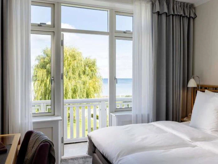 Et lyst hotelværelse med en pænt redt seng vender ud mod store vinduer og en balkon med udsigt over træer og en fjern udsigt over havet.