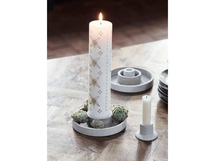 Et højt hvidt lys med guld snefnugdesign er tændt og placeret i en rund betonholder med små sukkulenter. Et mindre hvidt lys står ved siden af på et træbord.