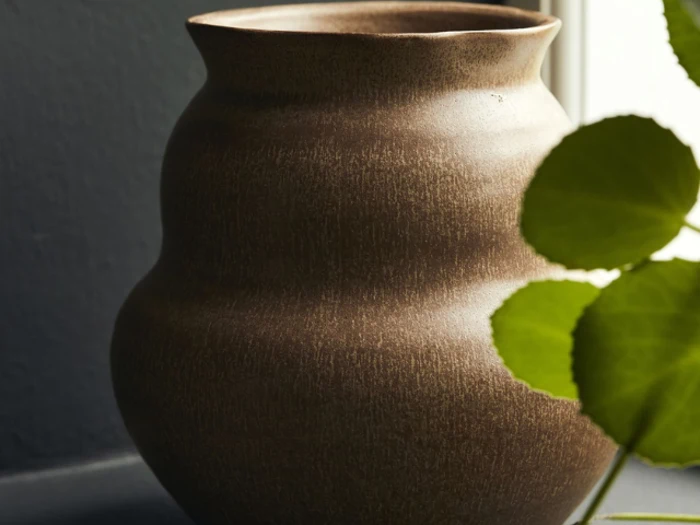 En brun, struktureret keramikvase står ved siden af et vindue, delvist skjult af grønne blade i forgrunden.