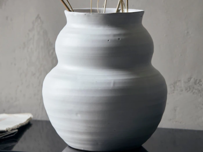 En hvid, rund, riflet keramikvase med en bølget silhuet, der holder et par tynde, høje pinde, står på en sort reflekterende overflade mod en grå struktureret væg.