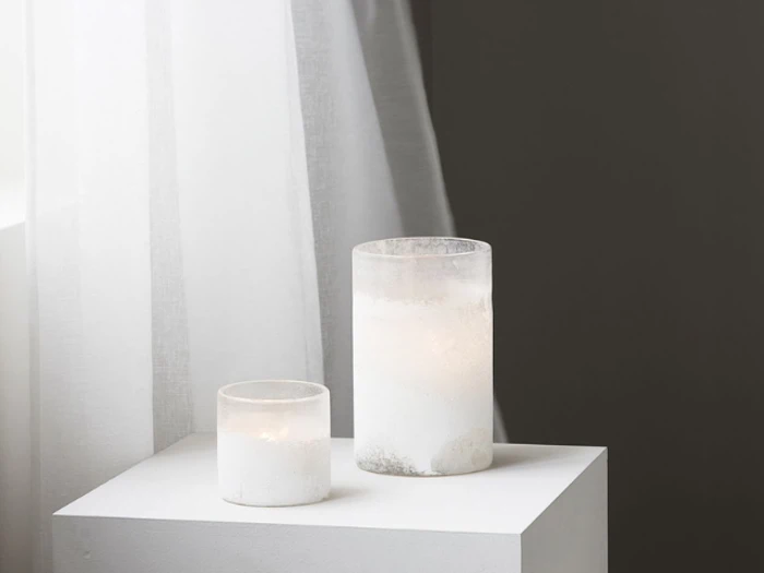 To hvide, matterede glaslys, et kort og et højt, står på en hvid piedestal ved siden af hvide, gennemsigtige gardiner på en mørkegrå væg.