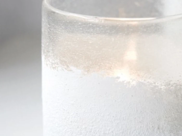 Et nærbillede af et frostet glas med koldt vand, hvor der dannes kondens på ydersiden. Baggrunden er ude af fokus og bleg.