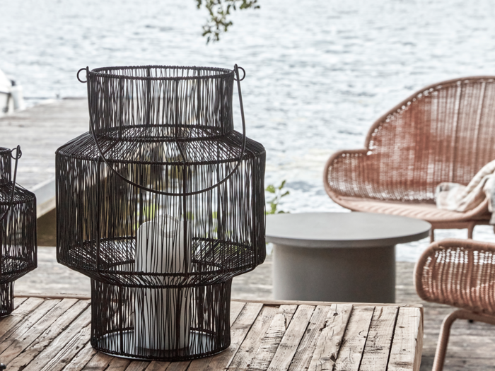 En sort trådlygte med et hvidt lys indeni står på et træbord udendørs i nærheden af kurvestole og en vandflade.