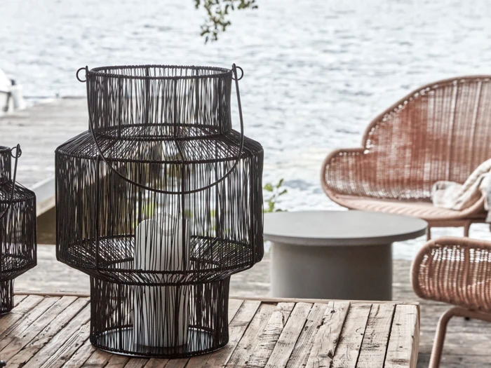 En sort trådlygte med et hvidt lys indeni står på et træbord udendørs i nærheden af kurvestole og en vandflade.