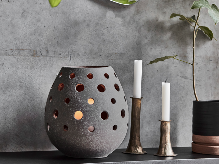 En grå, rund lanterne med runde udskæringer og et glødende lys indeni står ved siden af to hvide lys i metalholdere og en potteplante, alt sammen på en mørk hylde op ad en betonvæg.