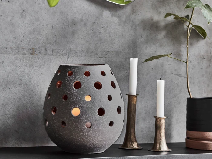 En grå, rund lanterne med runde udskæringer og et glødende lys indeni står ved siden af to hvide lys i metalholdere og en potteplante, alt sammen på en mørk hylde op ad en betonvæg.