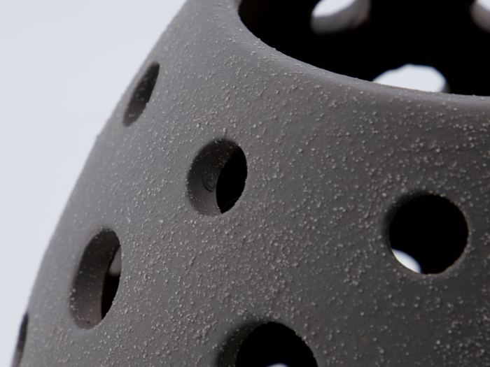 Nærbillede af en mørk, struktureret keramikgenstand med jævnt fordelte runde huller på den buede overflade.