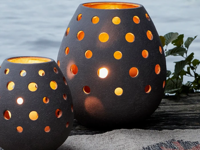 To sorte lanterner med cirkulære udskæringer lyser op i et varmt lys, mens de står udendørs på en træplade nær vand og grønne blade.