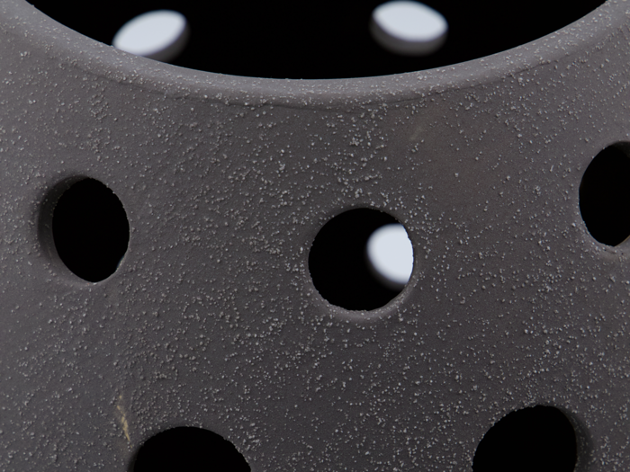Nærbillede af et struktureret, mørkegråt objekt med runde huller og en ru, plettet overflade. Hvidt lys er synligt gennem hullerne i baggrunden.