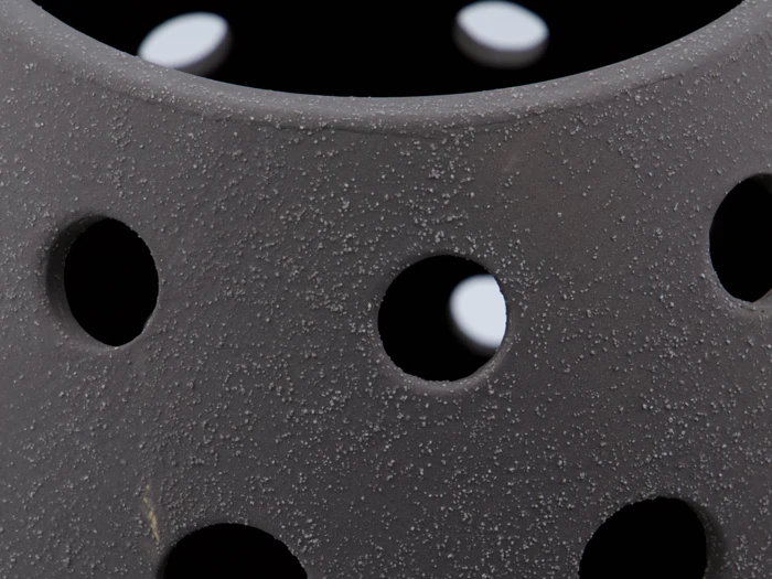 Nærbillede af et struktureret, mørkegråt objekt med runde huller og en ru, plettet overflade. Hvidt lys er synligt gennem hullerne i baggrunden.